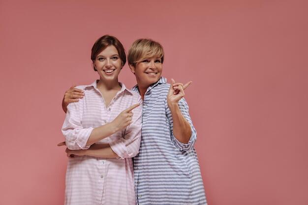 Free photo girl in striped pink dress smiling showing to place for text and hugging with old blonde woman in blue outfit on isolated backdrop